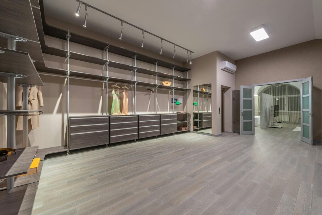 Stylish dresses and coat hanging on hangers in spacious walk in closet with empty shelves and opened doors in modern apartment